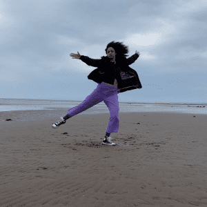 A person dancing on the beach wearing a black jacket and purple trousers.