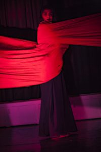 "Person looking into the camera with a red fabric draped around them. The fabric extends to either sides as if binding the dancer and restricting them. Still from the productions “What Draupadi Said to Penelopy” - by Dr Agomoni Ganguli Mitra