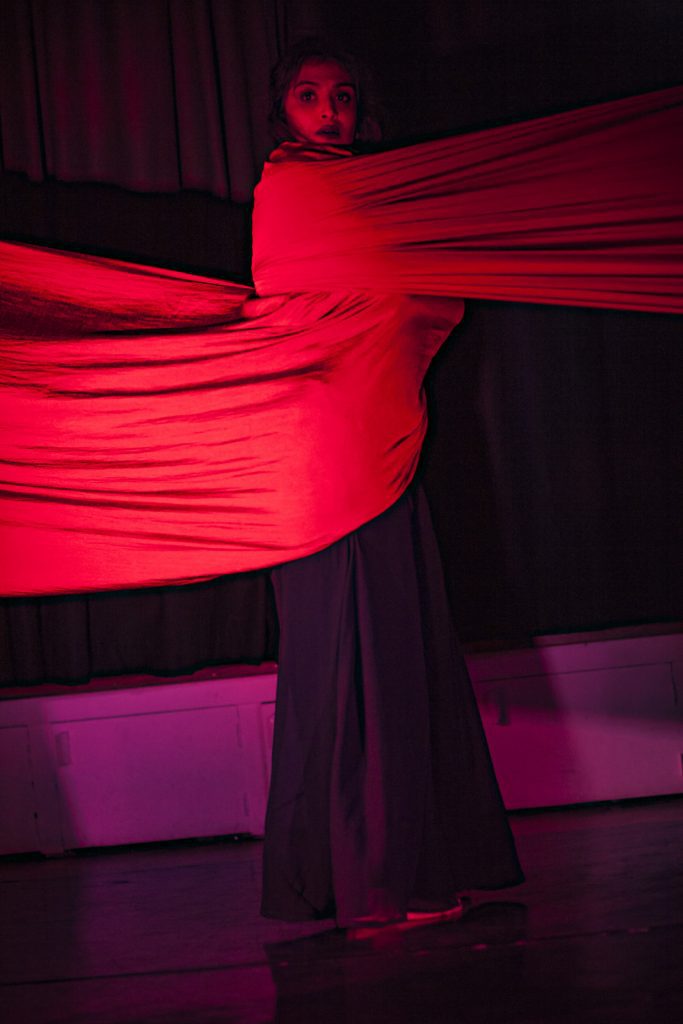 "Person looking into the camera with a red fabric draped around them. The fabric extends to either sides as if binding the dancer and restricting them. Still from the productions “What Draupadi Said to Penelopy” - by Dr Agomoni Ganguli Mitra
