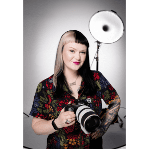 A photo of Lydia, who has black hair with a blonde streak and is standing in the centre of the image, holding a professional camera. She is standing in front of a white backdrop and there is photographic lighting equipment behind her.