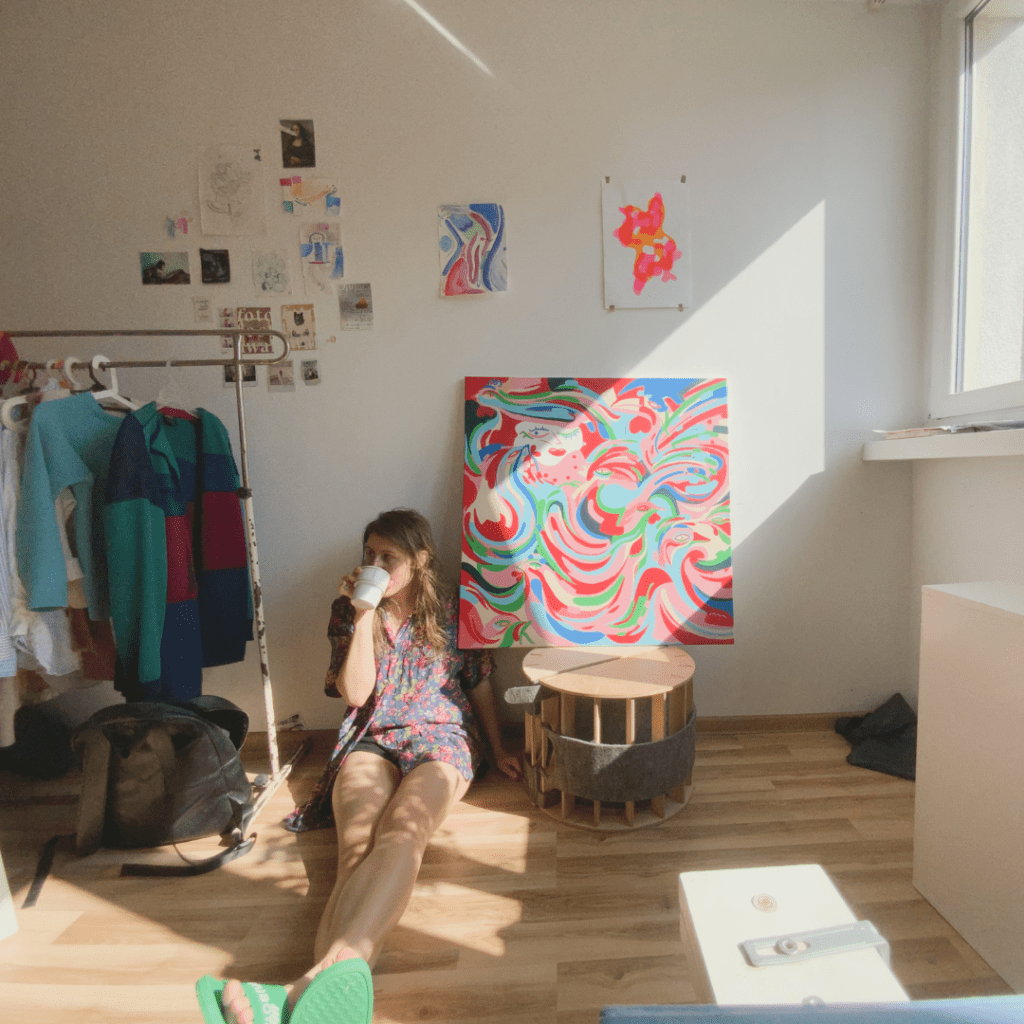 The artist sits on the floor of her studio next to a rail of clothes and a colourful painting.