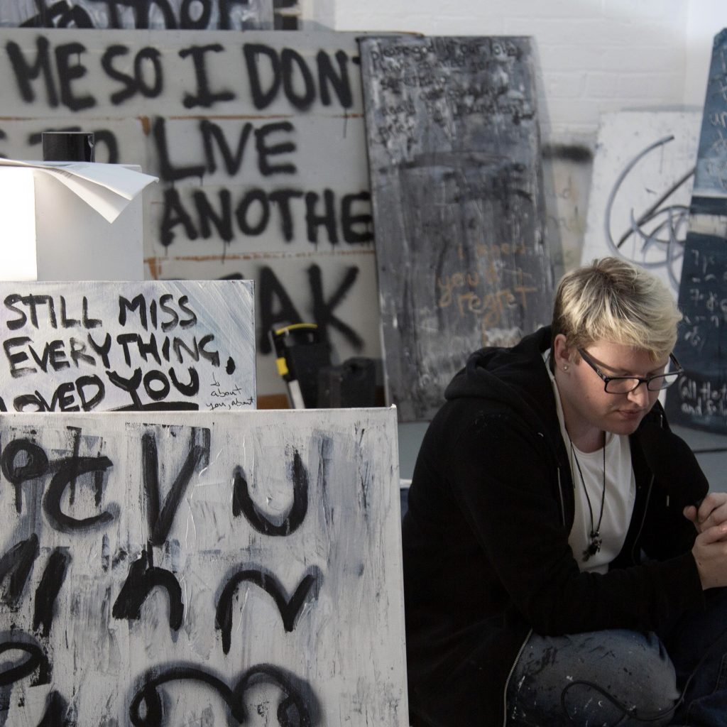 The artist - a white trans masc person with short bleached blonde hair - sits on a gallery floor surrounded by text-based paintings. Various words can be made out like “miss you” and “heartbreak”.