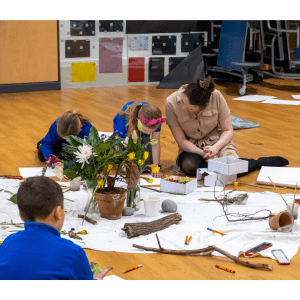 The artist sits on the floor with three children. They are working on a project, there are pens and paper on the floor in front of them.