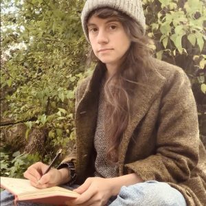 A white woman with long wavy brown hair sitting crossed-legged outside, with a pencil and sketchbook on her lap. It is Autumn, and she wears jeans, a thick green coat and a grey woolly hat.