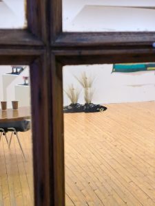 View through window pane into an exhibition space, with a distorted view of a sculpture made of earth, stones and grass.