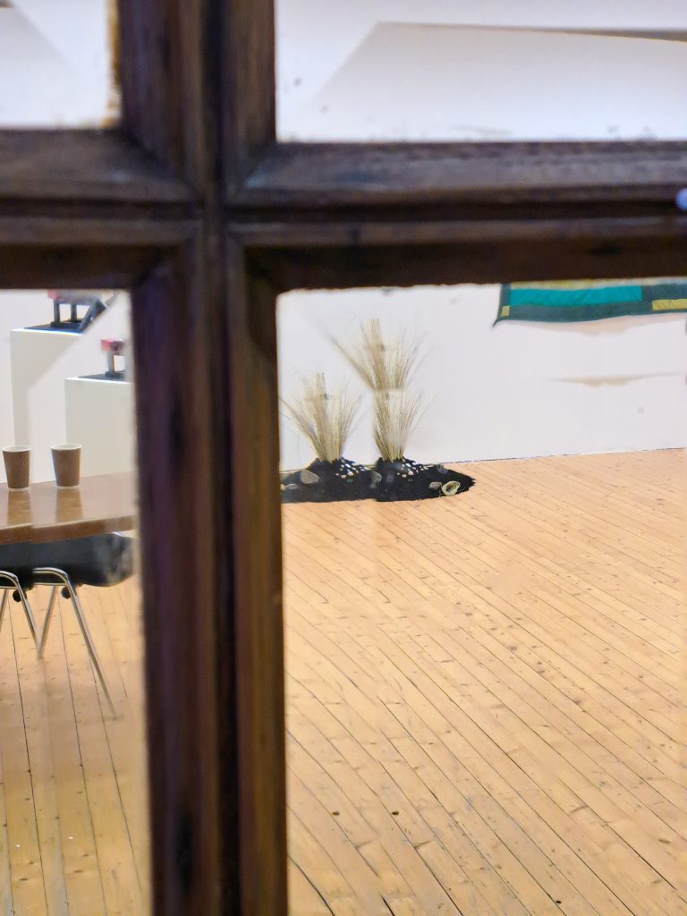 View through window pane into an exhibition space, with a distorted view of a sculpture made of earth, stones and grass.