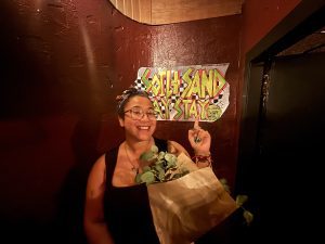 Hannah stands in front of a red wall, dressed in black and holding a bouquet of eucalyptus. Behind her head on the red wall is a sign reading ‘Soil And Sand Say Stay - exhibition August 2025’