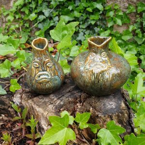 Two witch bottles, one smaller and one larger and more round, in a garden setting. Ivy covers the background. The bottles are ceramic earthenware, with a base colour of warm brown and, on top, a forest green drip. Each bottle has a frowning face near the neck.