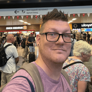 A white, male presenting figure in a train station, wearing glasses and a pink t-shirt.