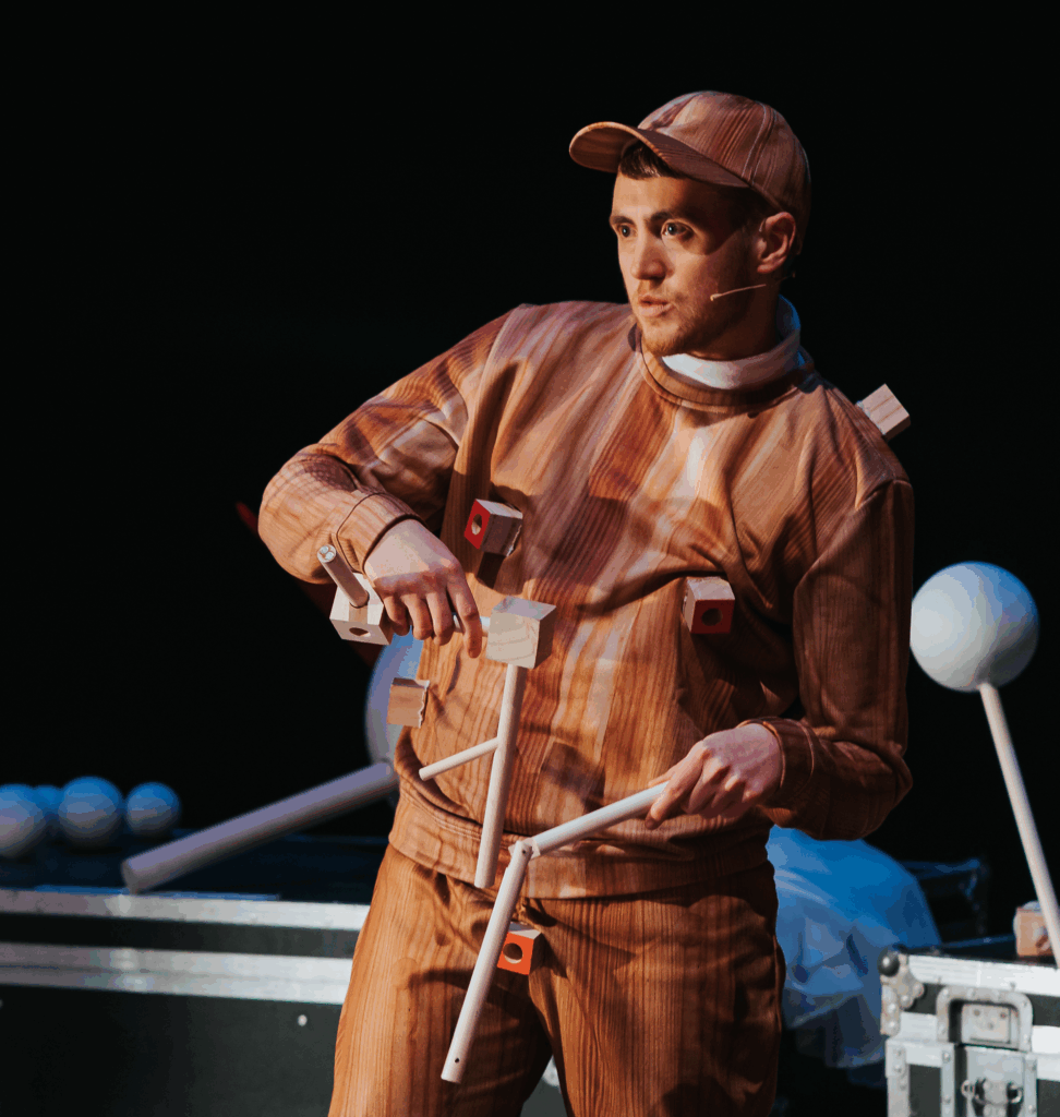 An image of Ivor performing in The Making of Pinocchio. They are a white trans man with a short gingery beard. They wear a cap and tracksuit that has a fake wood pattern, and has wooden blocks sticking out of it at the nipples, shoulder and groin. Ivor is holding two wooden objects made of wooden dowels and blocks and he has a little microphone on his face.