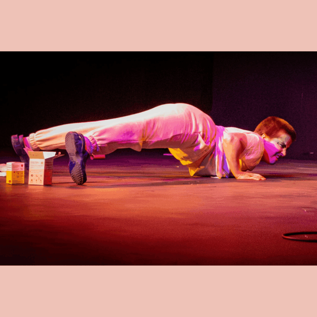 The image shows Narcisse Empoté-Ose caught in a moving plank on stage. He is wearing white pijamas, jogger style, and blue fake Crocs. His face has traditional drag king makeup (beard and moustache) and Narcisse's expression is showing effort. The light is stage light, very red and bright. On the floor around Narcisse, there are period product boxes.