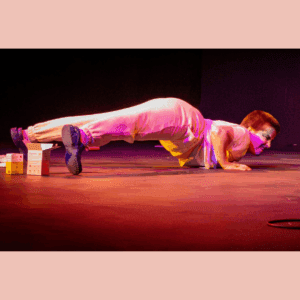 The image shows Narcisse Empoté-Ose caught in a moving plank on stage. He is wearing white pijamas, jogger style, and blue fake Crocs. His face has traditional drag king makeup (beard and moustache) and Narcisse's expression is showing effort. The light is stage light, very red and bright. On the floor around Narcisse, there are period product boxes.