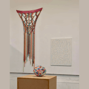 Photo of an exhibition. There is a sculpture on a plinth in the foreground, it looks like a very large ball of rubber bands. In the background there is a colourful textile piece hanging on the wall next to a white canvas with black markings.
