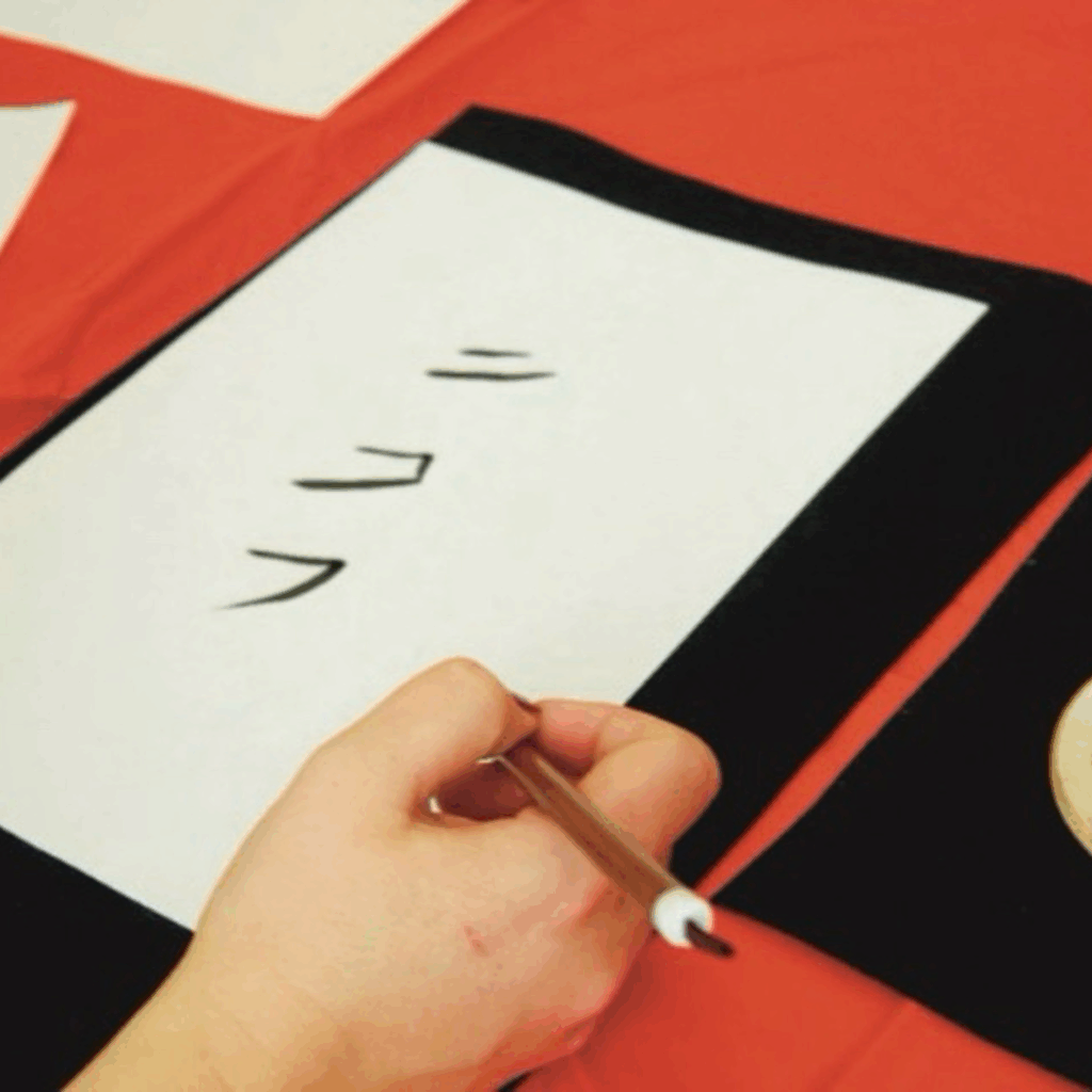 A hand holding a paintbrush is poised over a piece of paper with Japanese calligraphy on it. The background shows a red tablecloth.