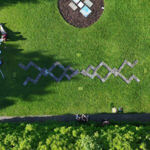 A piece of land art, taken from above. Two trellis-like shapes are laid on the grass, in a garden. There is a circular flowerbed at the top of the frame that has 4 pieces of paper lying in it. The shadow of a tree almost obscures one of the trellis shapes.