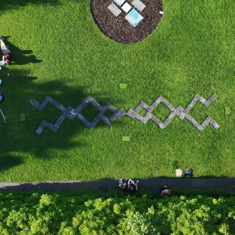 A piece of land art, taken from above. Two trellis-like shapes are laid on the grass, in a garden. There is a circular flowerbed at the top of the frame that has 4 pieces of paper lying in it. The shadow of a tree almost obscures one of the trellis shapes.