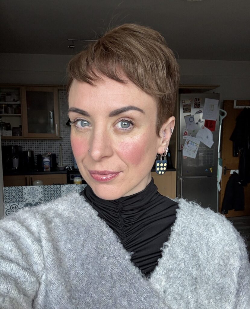 A portrait photograph of a woman smiling wearing a fuzzy grey jumper and a black polo neck.
