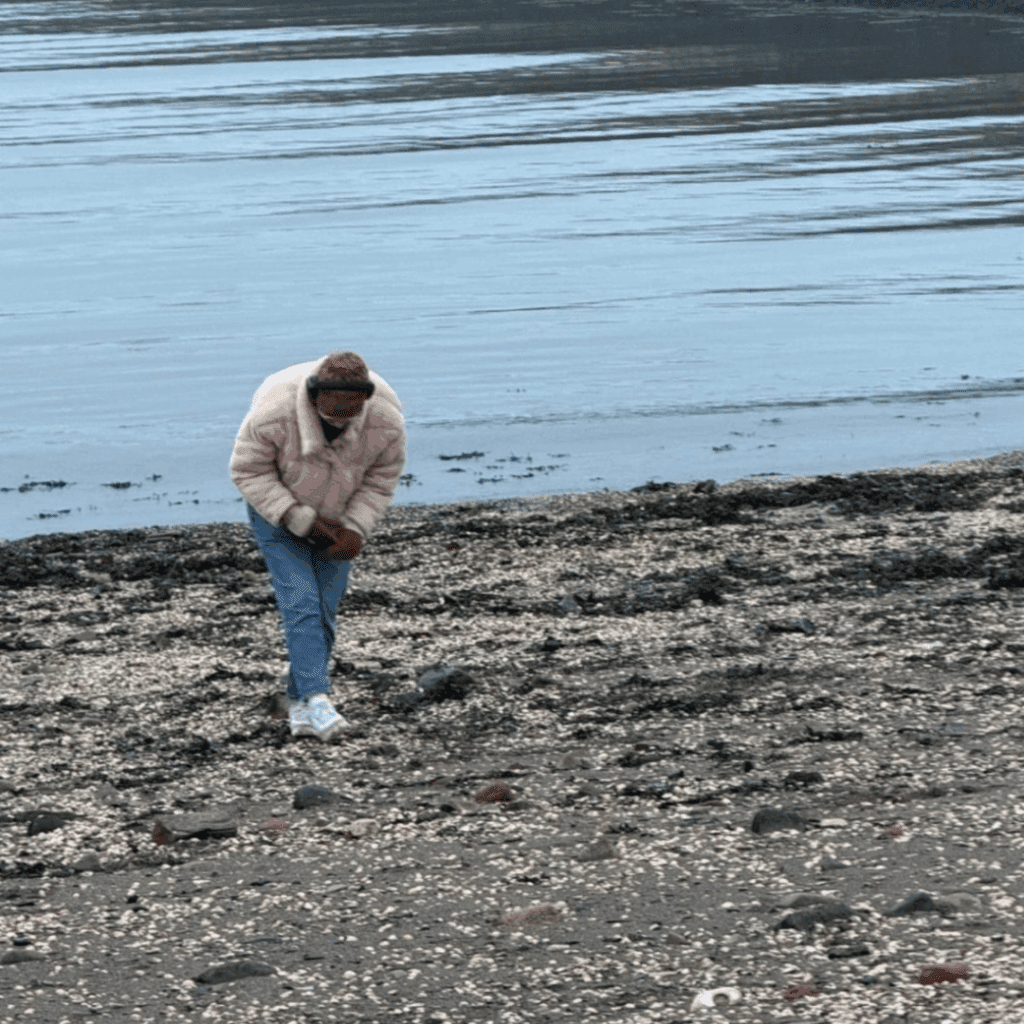 A picture of someone walking, bent over, on a stony beach. The water is still and calm. They are wearing a pink jacket and blue jeans and appear to be looking for something.