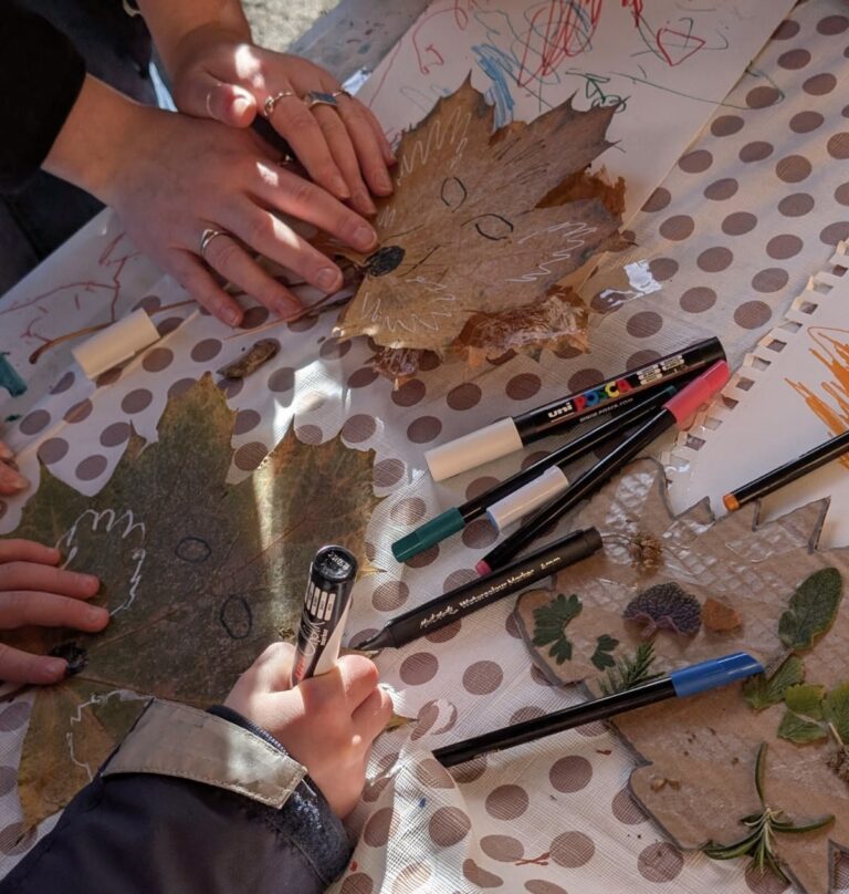 The image shows a table covered in a floral table cloth with art materials, books and leaves strewn across it. On top, there are a pair of adult's hands and a pair of children's hands decorating a large pressed leaf to look like a fox.