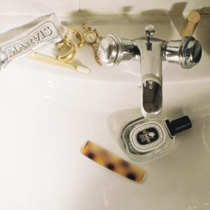 A film photograph of a bathroom sink cluttered with a toothbrush, toothpaste, lipstick, jewellery, a perfume bottle and a comb.
