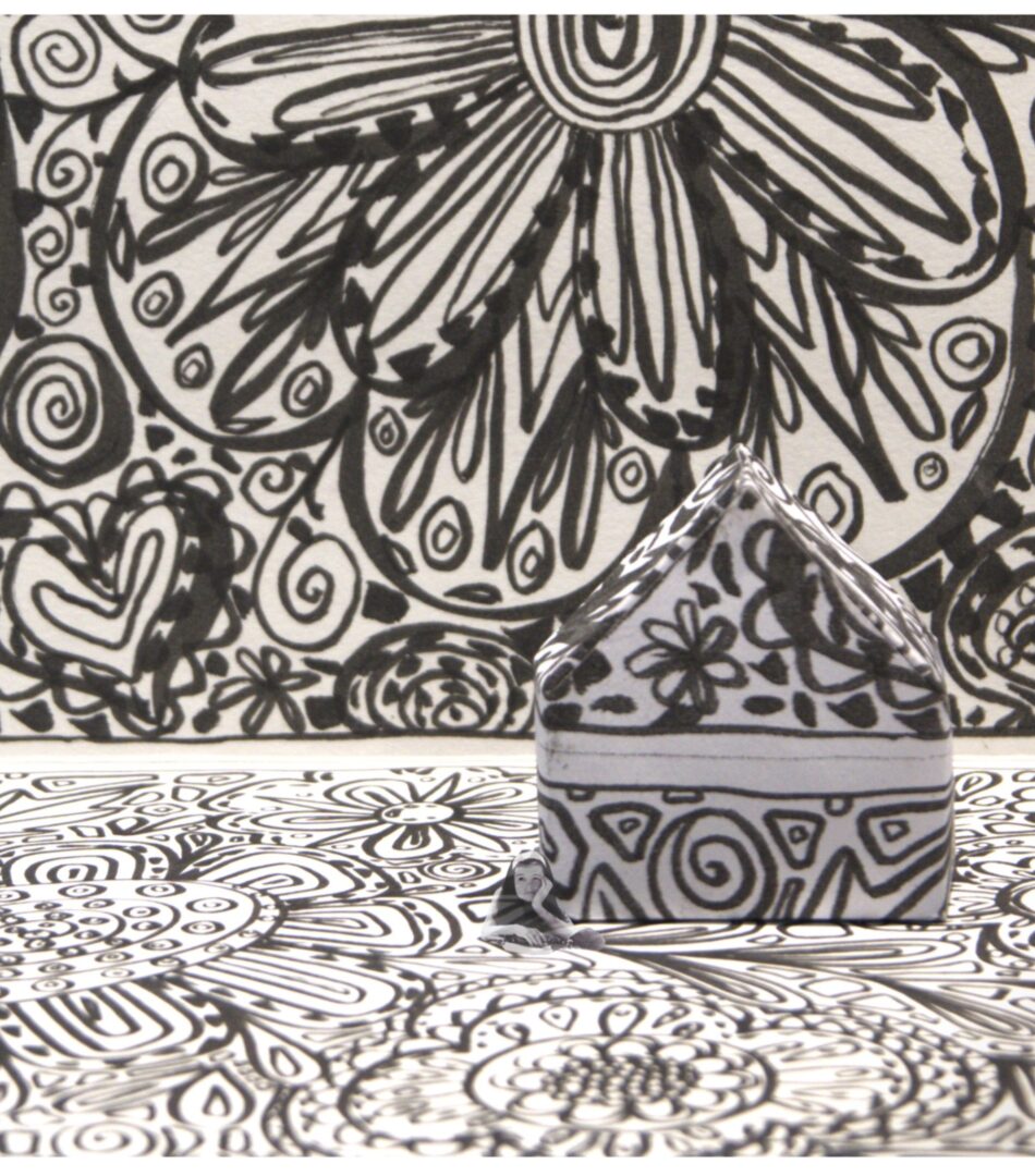 Black-and-white photo of a patterned paper house against a matching patterned background, with a tiny vintage photo of a person in front.