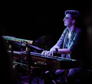 Lesley, a slim built, tall woman with short grey-black hair, wearing glasses sits behind two keyboards on a double stand. She is playing the lower keyboard.