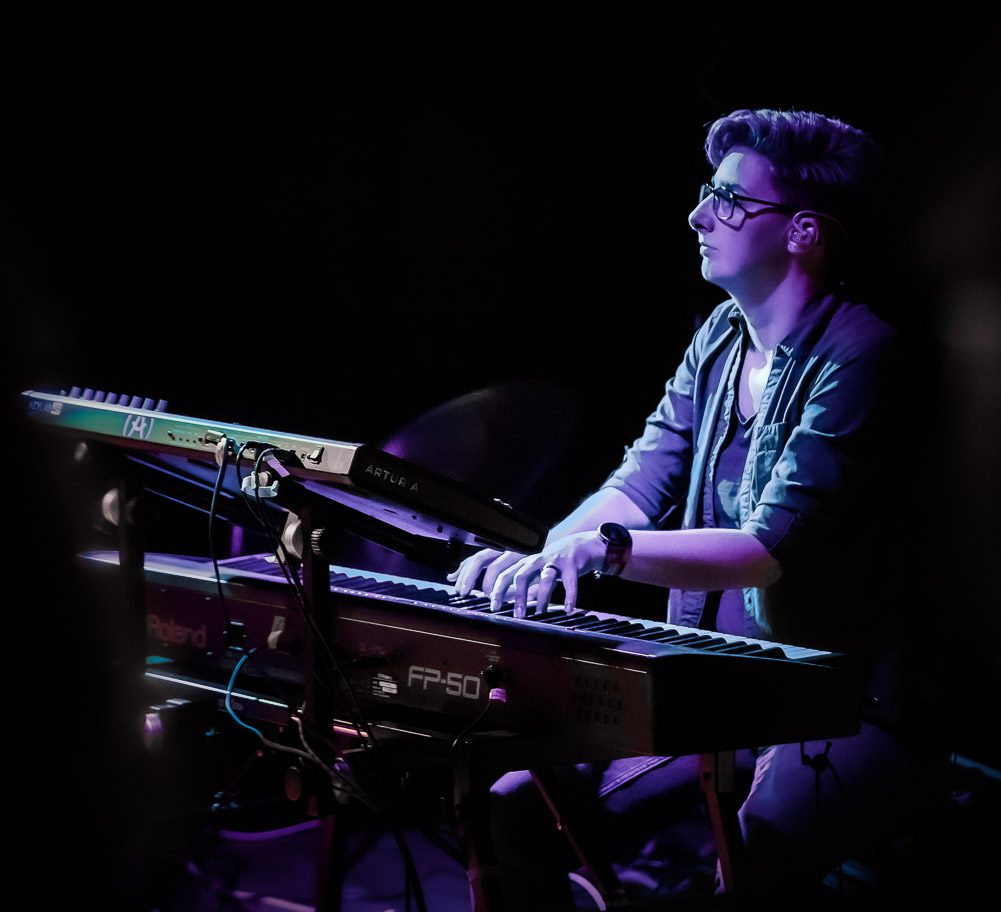 Lesley, a slim built, tall woman with short grey-black hair, wearing glasses sits behind two keyboards on a double stand. She is playing the lower keyboard.