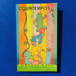 Photograph of a magazine with illustrated cover of a white character with big nose standing in tall grass. They wear a tower of multicoloured hats stacked atop their head. A worm threads it's way between the hats. The characters hold a very long bendy paintbrush and a long grabber, rearranging and painting the letters above that read "COUNTERPOINT". Multicoloured striped wallpaper is behind them.