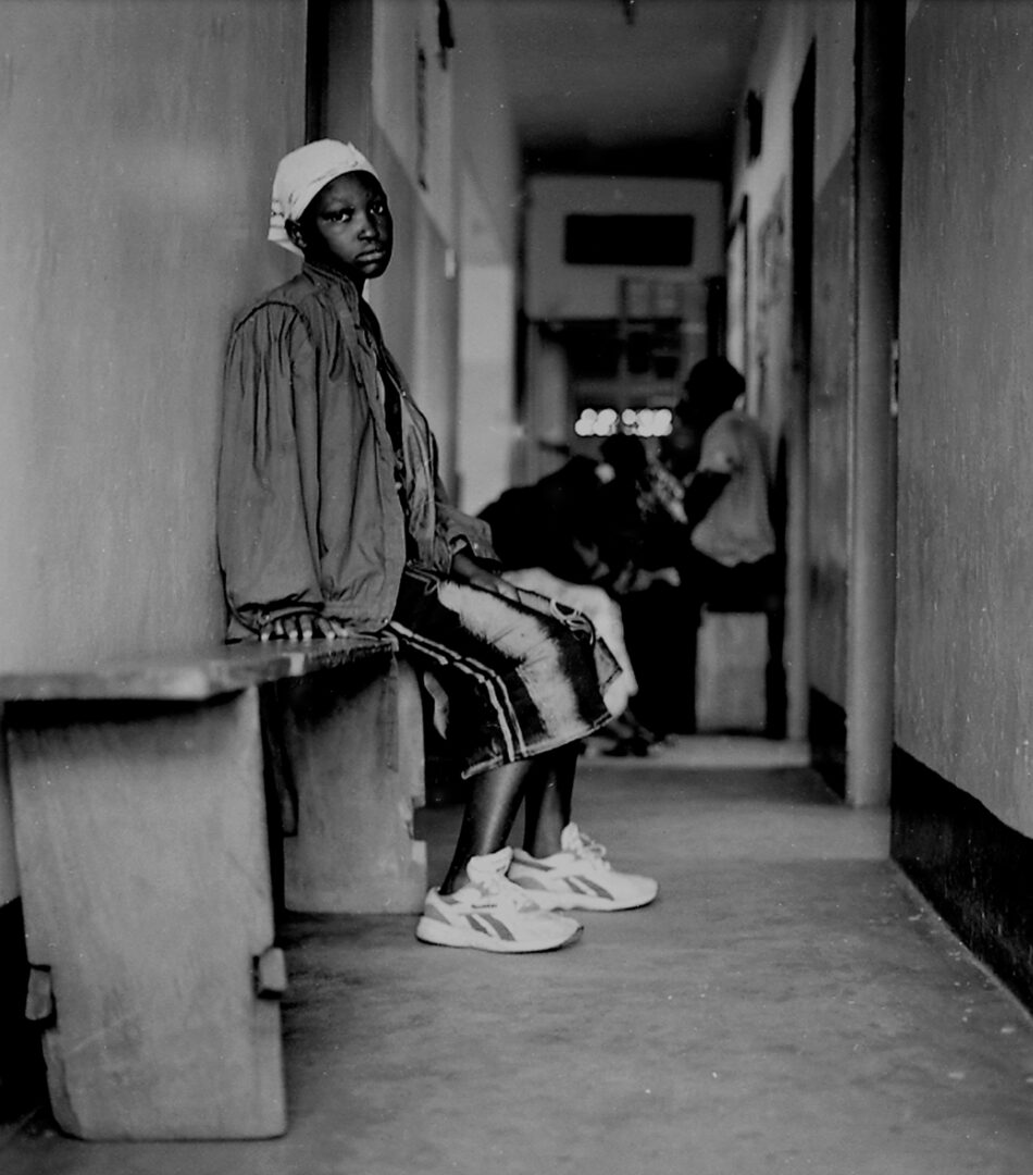 A black-and-white study of a figure in a health clinic. The subject sits on a heavy, wooden bench on the left, their gaze directed toward the camera with a steady, quiet presence. A low-angle perspective, which grounds the viewer on the floor of the corridor and emphasises the physical weight of the environment.