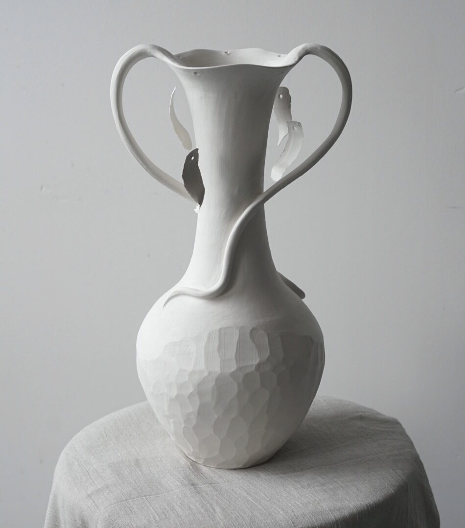White biscuit fired vase with a faceted bottom and a tall neck which opens into a more floral shape. Shards from the faceted bases are hung from the mouth of the vessel.