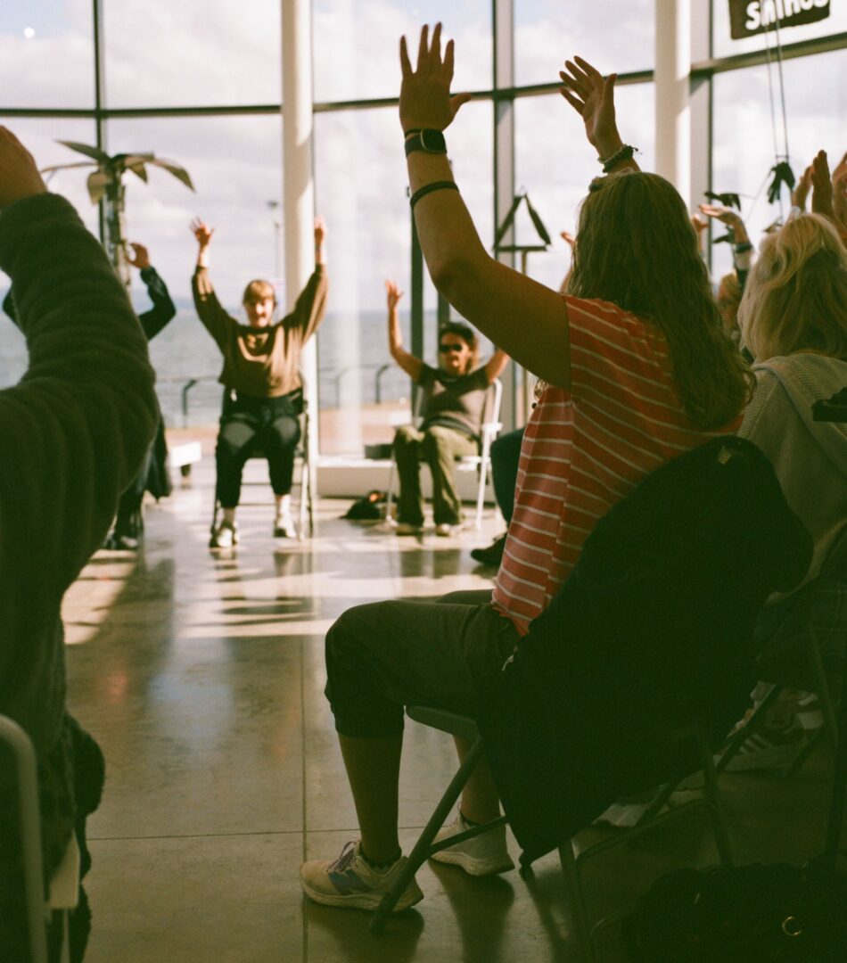 A group of women sitting on chairs in a circle with their arms raised in the air. Behind them are large windows.