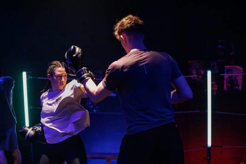 Two people spar in a boxing ring under colourful neon lighting; a woman in a white top throws a punch while her male partner blocks.