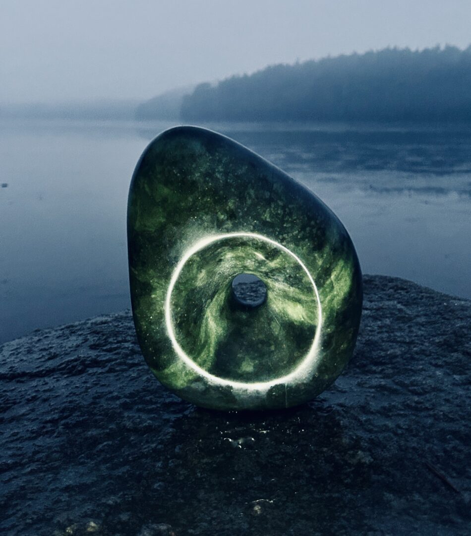 A carved turquoise stone placed on a rock with water in the background. There is a glimmer of circular light in the carved design of the stone, which contrasts against the dark background.