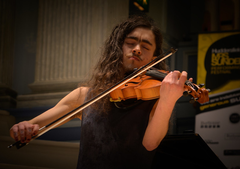 Eldon, a white, thin, androgynous person with long brown hair, plays the viola with their eyes close. They are wearing a silvery blue dress, and only their top half in visible.