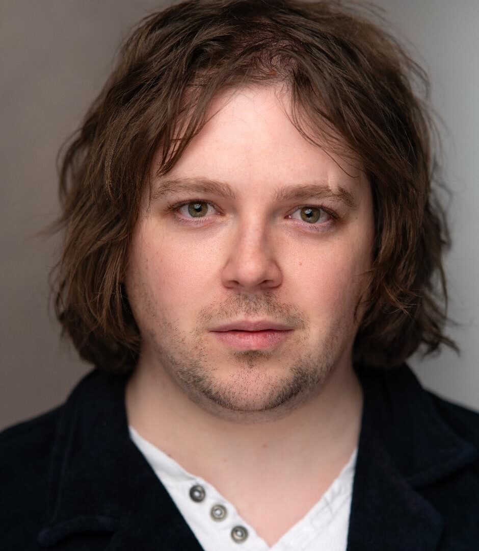 A headshot of Ell wearing a white top and blue jacket. They have longish brown hair, white skin and short stubble. They also have a scar on one eyebrow, and they are slightly smirking.