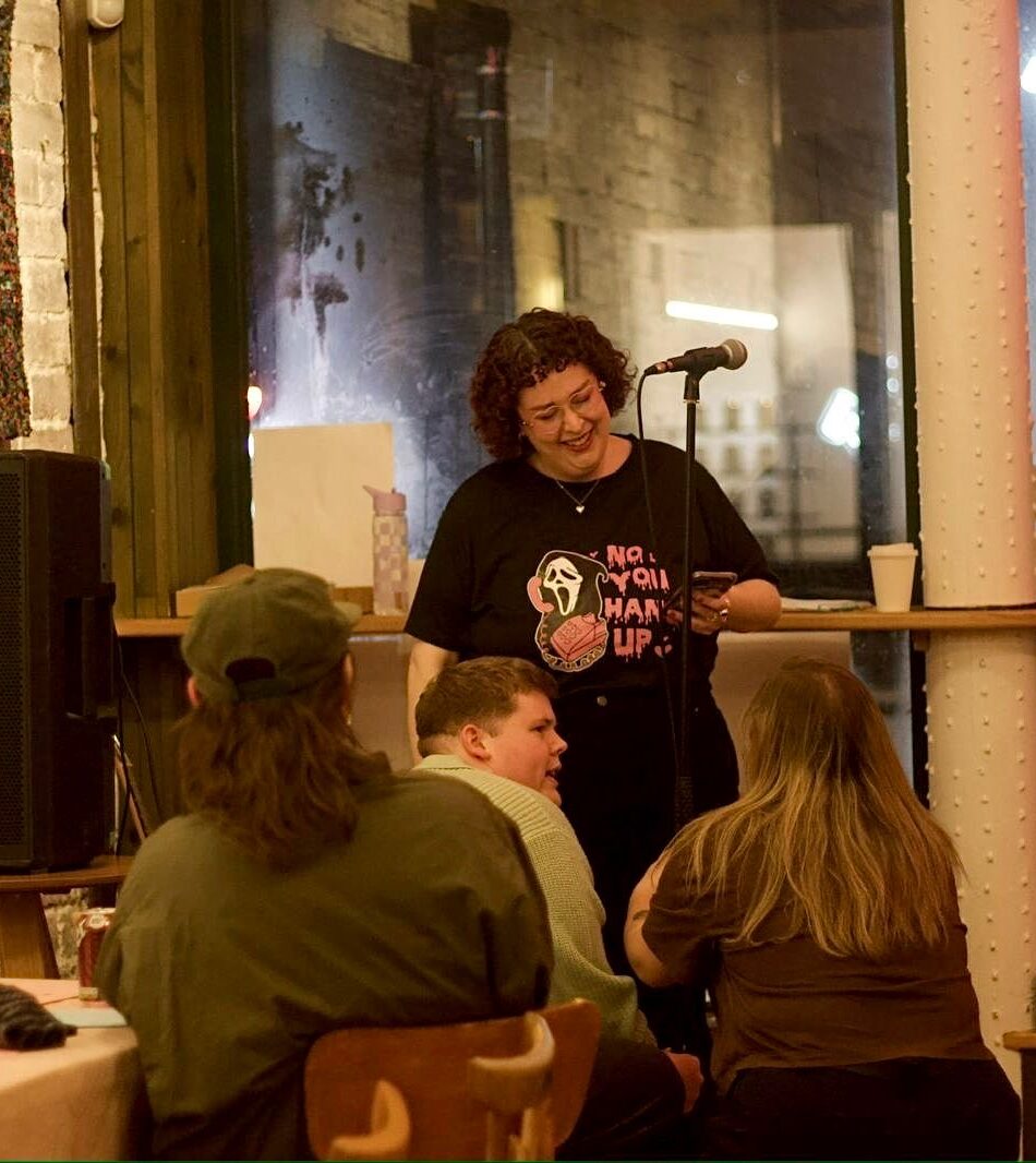 A white woman with short brown hair stands at the front of a room full of people, performing poetry.