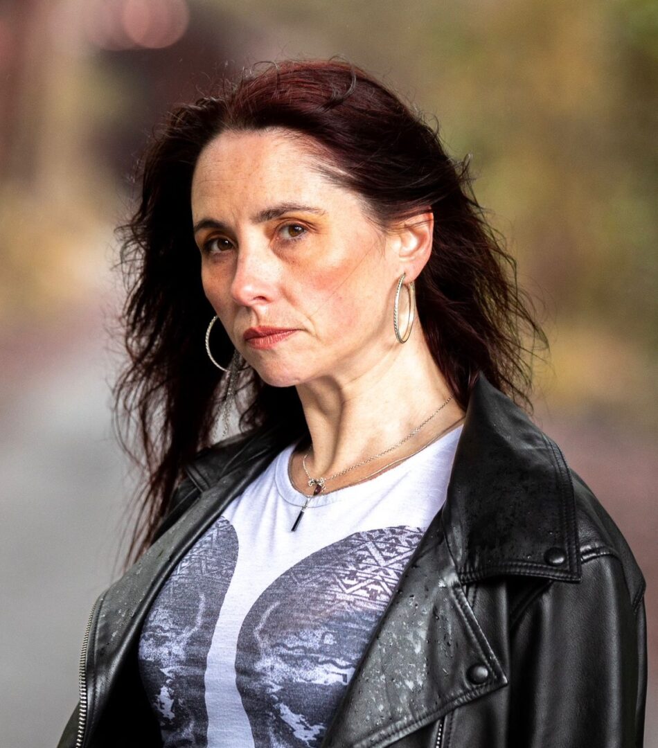 Headshot of a young white woman with brown hair, wearing a T-shirt and a leather jacket. She looks at the camera defiantly.