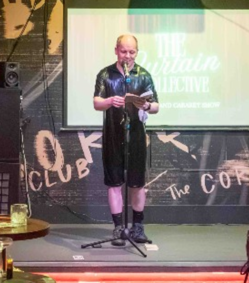 Thie photo depicts me on stage wearing a black PVC bodysuit, black trainers and socks and a white facemask around my left wrist, reading a poem from my poetry book to an audience from the stage. The event is the Curtain Collective held at the Corset Club in Glasgow.