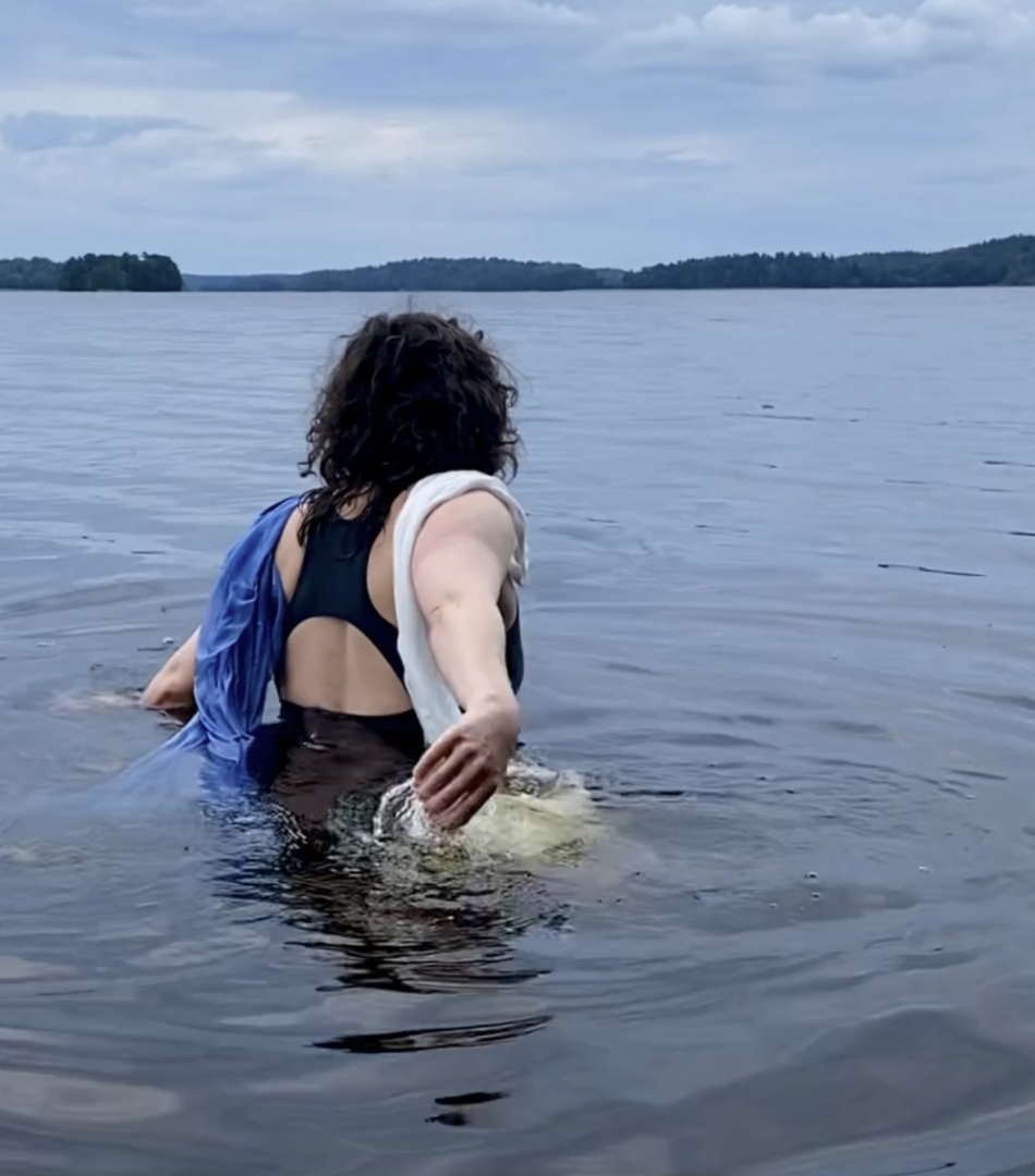 The artist is in water with her back turned moving to swim into it. Fabric is wrapped on her shoulders on a bright day.