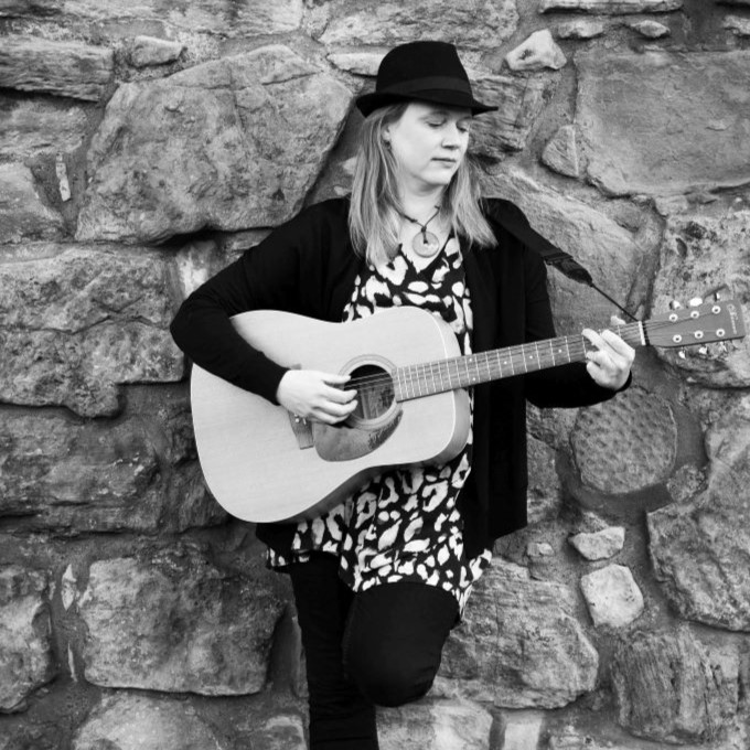 Black and white photo with artist standing with back to stone wall holding a guitar and looking down to the side.