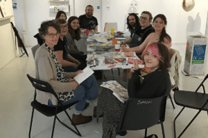 A large group of people sitting around a table covered in collage materials. They are looking at the camera and smiling