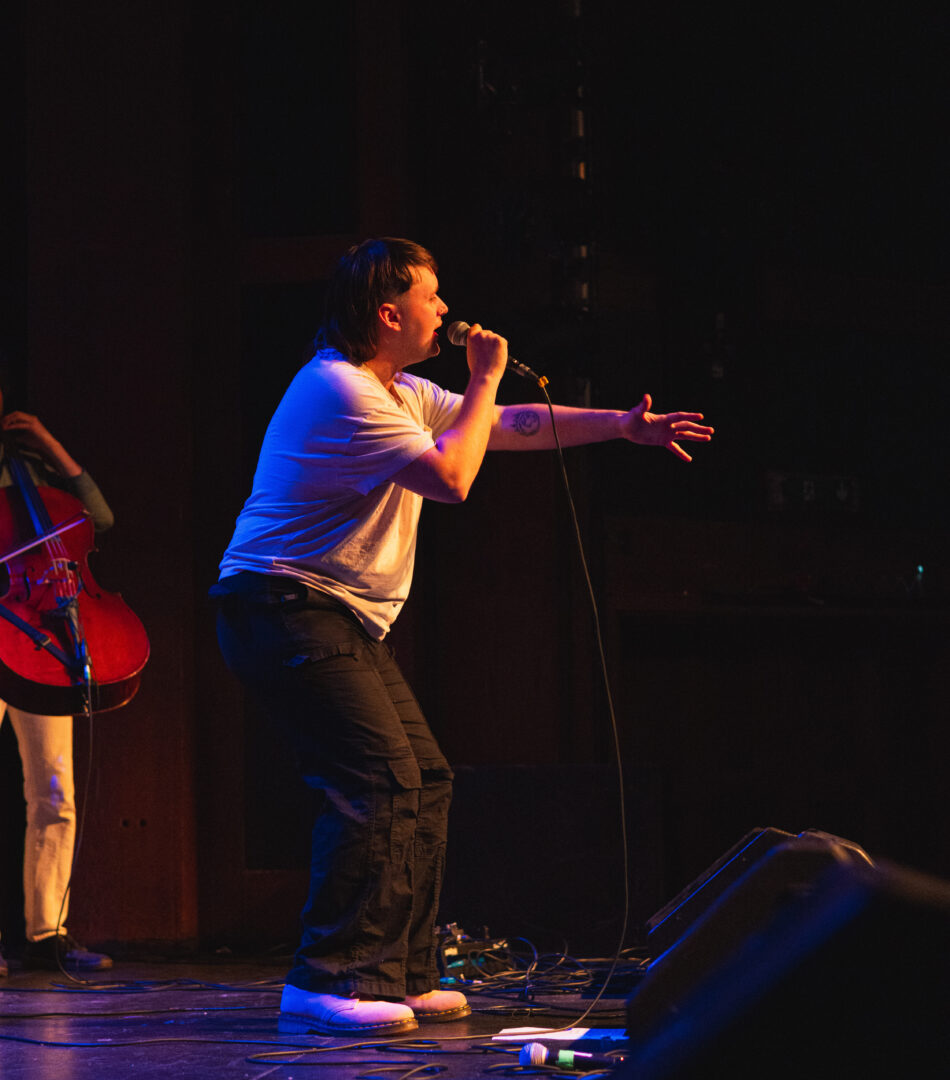 A non-binary individual wearing white t-shirt and black trousers holding a microphone and gesturing to an audience while performing with a live orchestra.