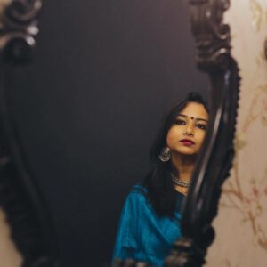 A young South Asian woman reflected in an ornate mirror. She is wearing a blue shawl and large, ornate silver earrings. She has a serious expression.