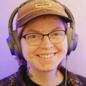 A smiling non-binary person wearing glasses, ear defenders, and a baseball cap.