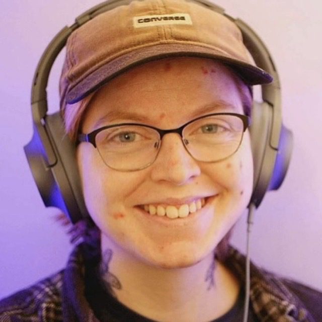 A smiling non-binary person wearing glasses, ear defenders, and a baseball cap.