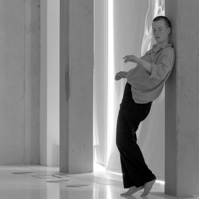 Black and white photo of a person with cropped hair leaning artistically against a wall - they seem almost to be mid-dance.
