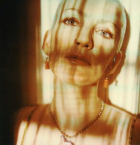 A woman with short blonde hair is lit by warm sunlight and striped shadows, looking upward with a calm expression.