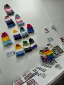 A white gallery wall of knitted hats hanging loosely, each a different pride flag. Three are on black stands on a table beneath the others, the rainbow flag, simplified lesbian and simplified gay men pride flags. Small pride flag stickers are also laid out on the table, the colours are not very discernable from the distance the picture was taken. Work from other artists is also visible on the wall and table.