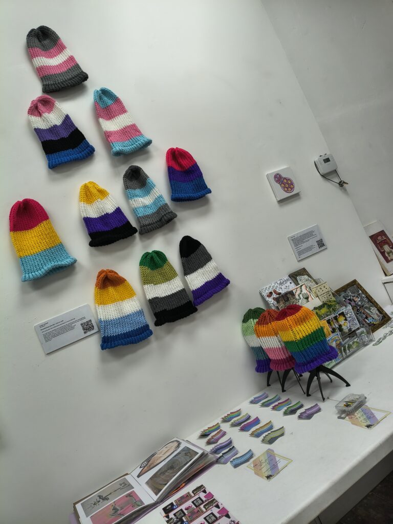 A white gallery wall of knitted hats hanging loosely, each a different pride flag. Three are on black stands on a table beneath the others, the rainbow flag, simplified lesbian and simplified gay men pride flags. Small pride flag stickers are also laid out on the table, the colours are not very discernable from the distance the picture was taken. Work from other artists is also visible on the wall and table.