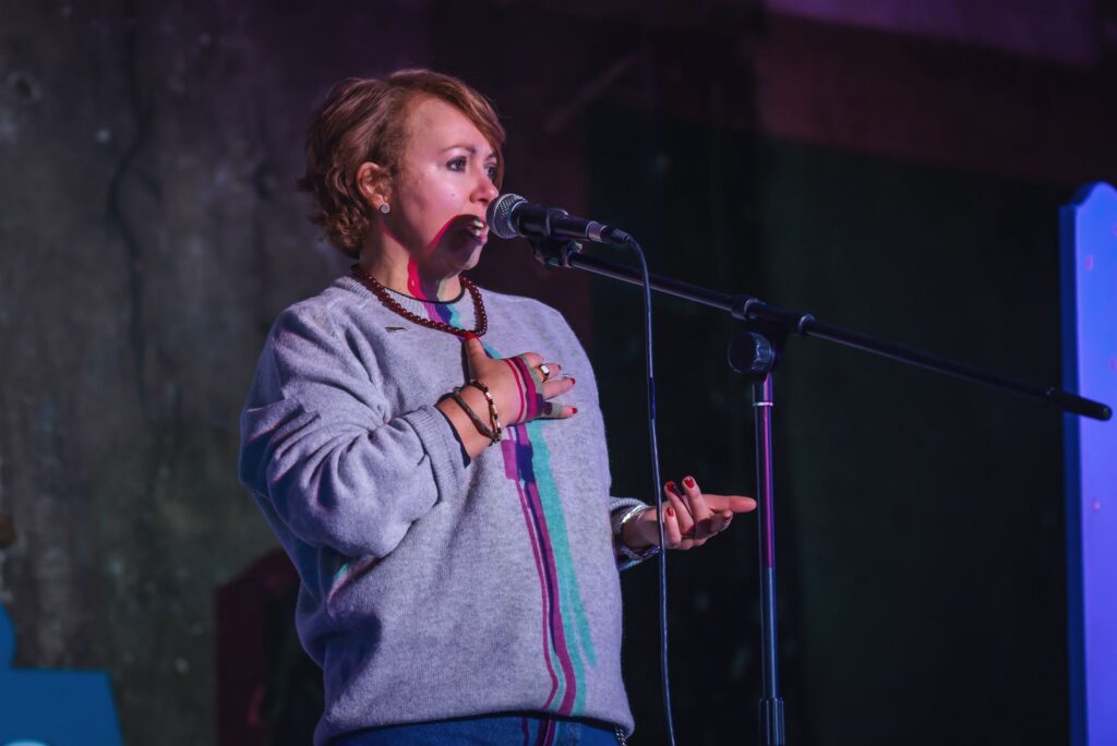 Cat Cochrane performs at the Scottish National Poetry Slam Championship at the Panopticon in Glasgow on Feb 14. Cat stands at the mic looking out to the audience. Cat has red hair and is wearing a grey jumper.